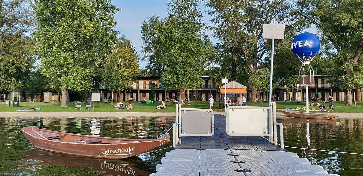 Zum Baden in Wien besucht die Klasse die Alte Donau. Auf dem Bild das Strandbad Gänsehäufel vom Wasser aus fotografiert. Im Vordergrund der Steg mit Boot, im Hintergrund die Badewiese.